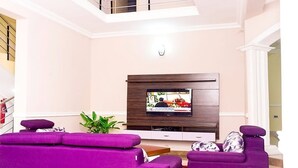 A modern living room with a flat-screen TV mounted on a wooden wall, purple sofas, and a staircase leading to an upper level. - The Good House PHC (Port Harcourt)
