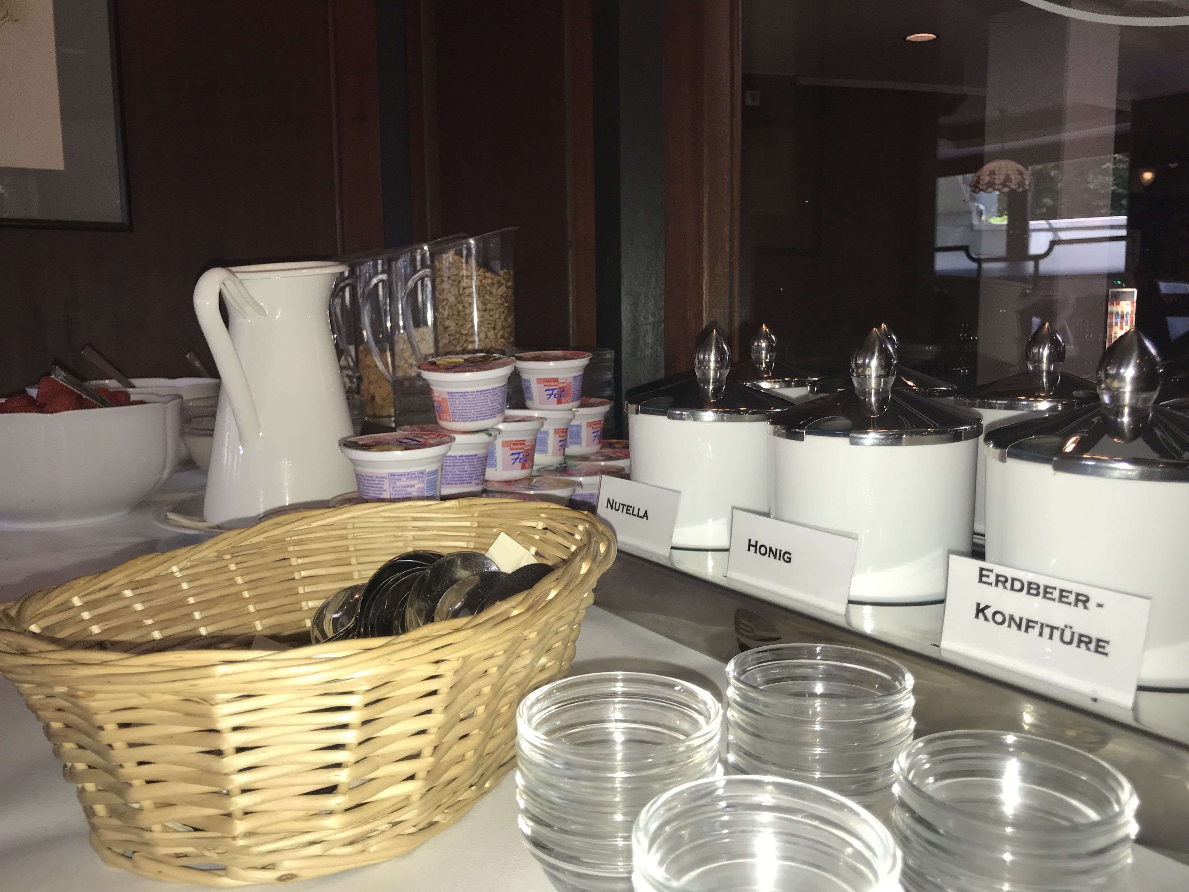 A buffet setup with labeled containers for Nutella, Honig, and Erdbeer-Konfitüre, a wicker basket, and clear plastic cups.