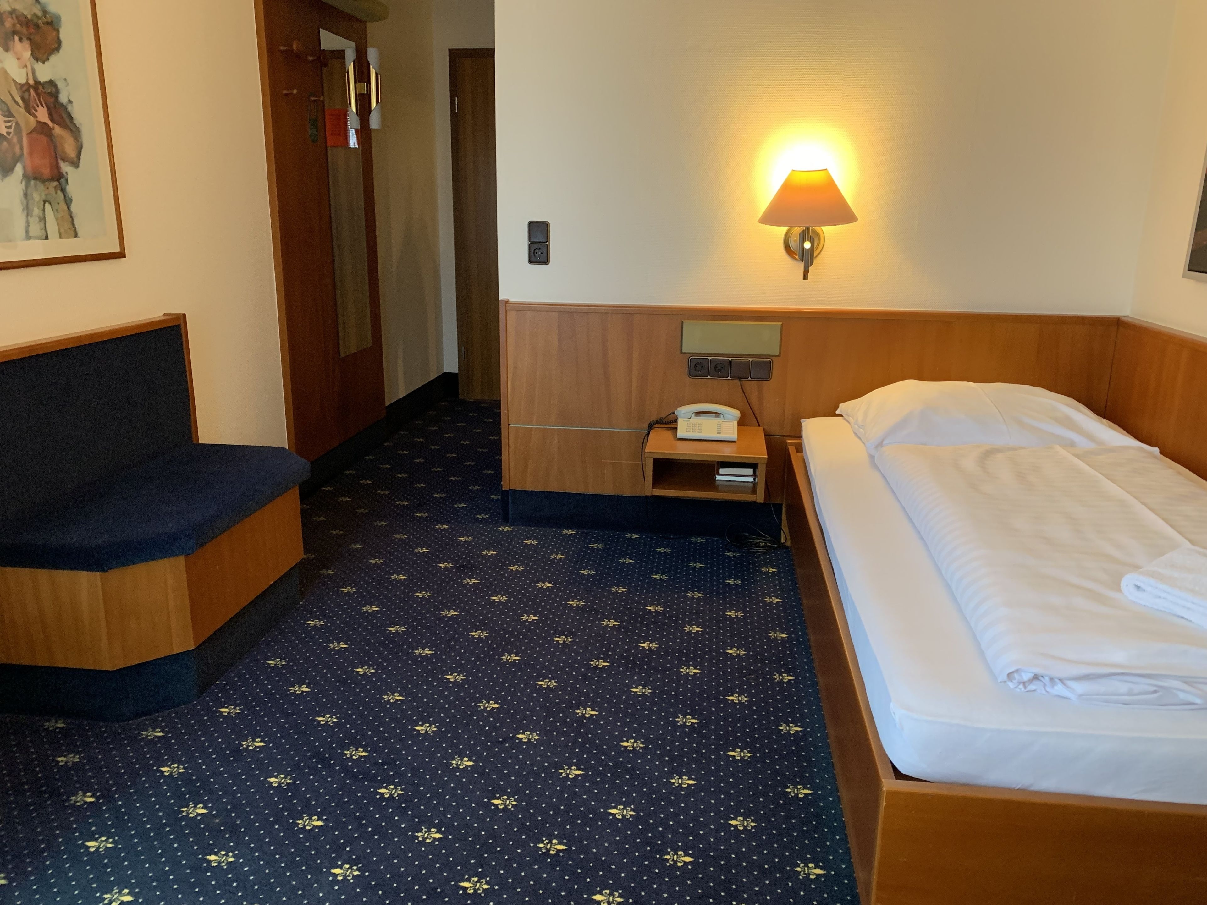 A hotel room with a bed, a desk with a lamp, a telephone, and a painting on the wall.