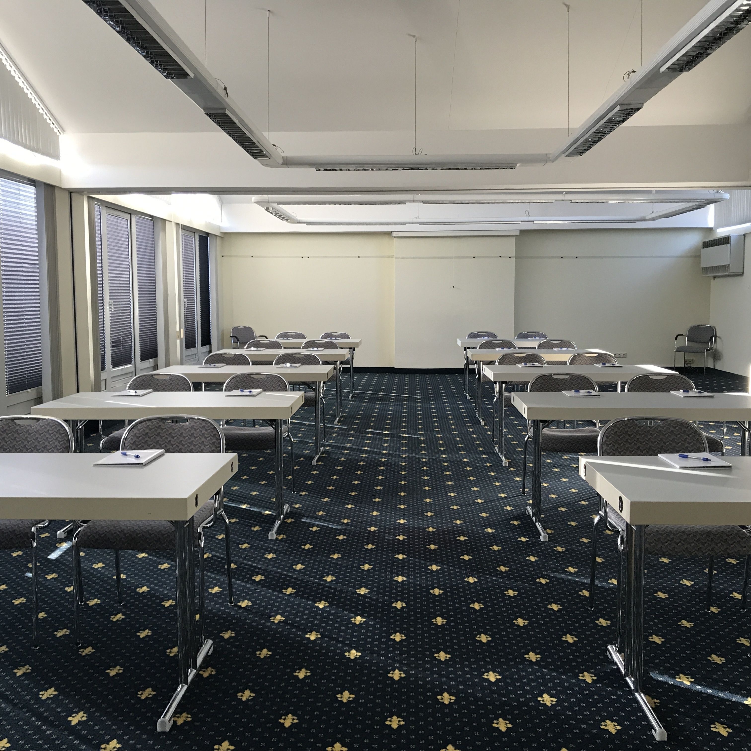 A conference room with tables and chairs, a patterned carpet, and a plain wall with an air conditioning unit.