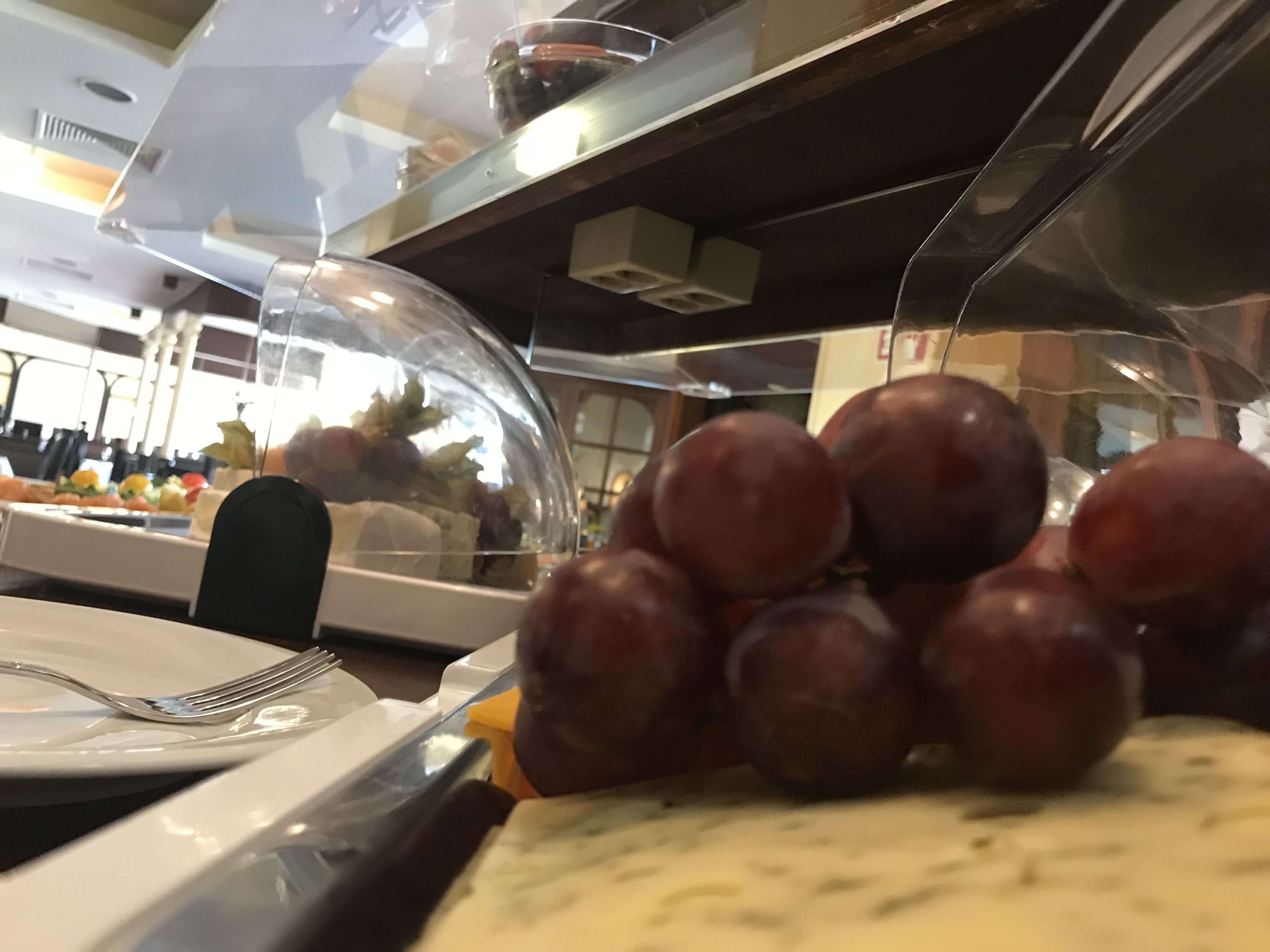 A buffet area with a glass dome covering food items, including a pile of grapes and a plate with a fork.