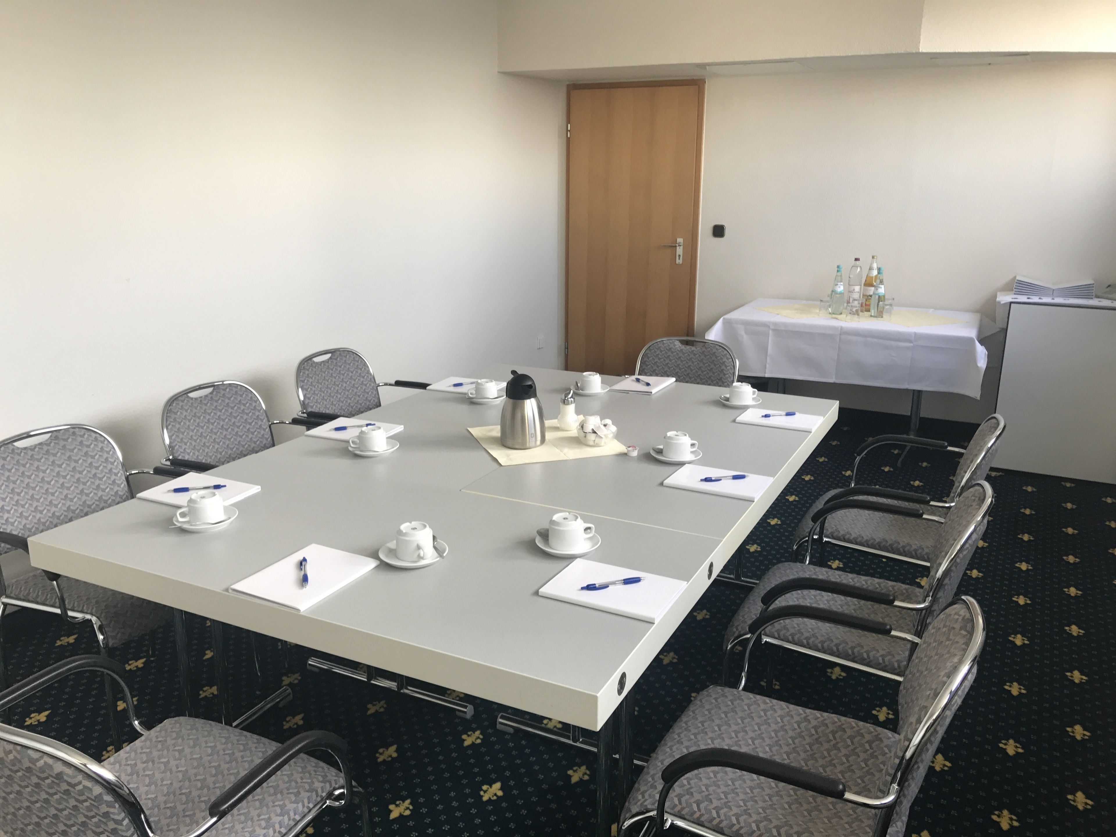 Un salón de conferencias con una larga mesa, sillas y una mesa pequeña con botellas y un mantel blanco.