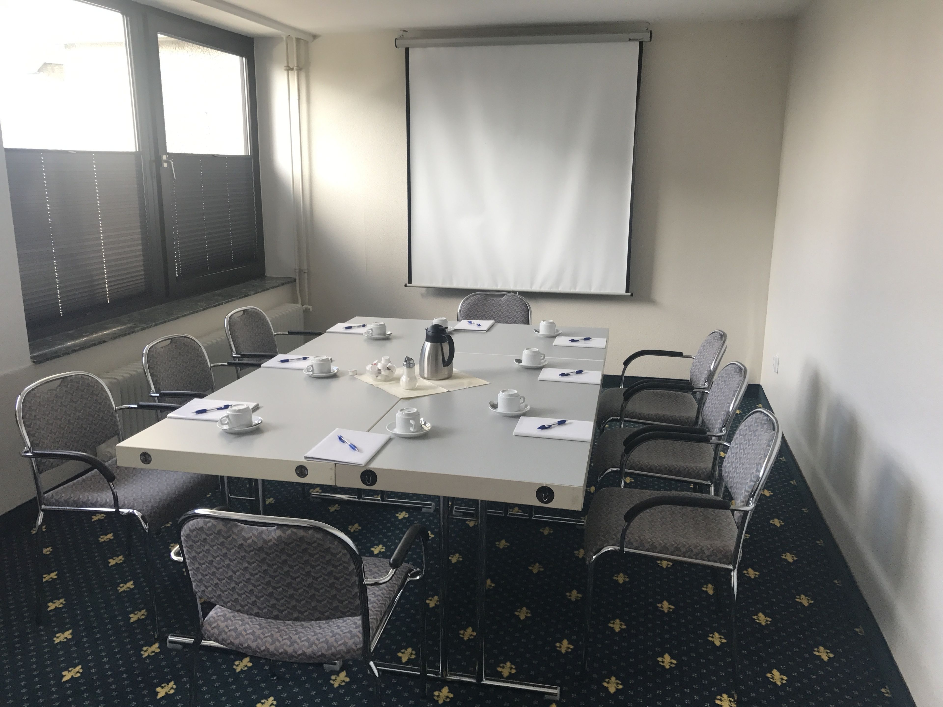 A conference room with a large table, chairs, a projector screen, and a window with blinds.