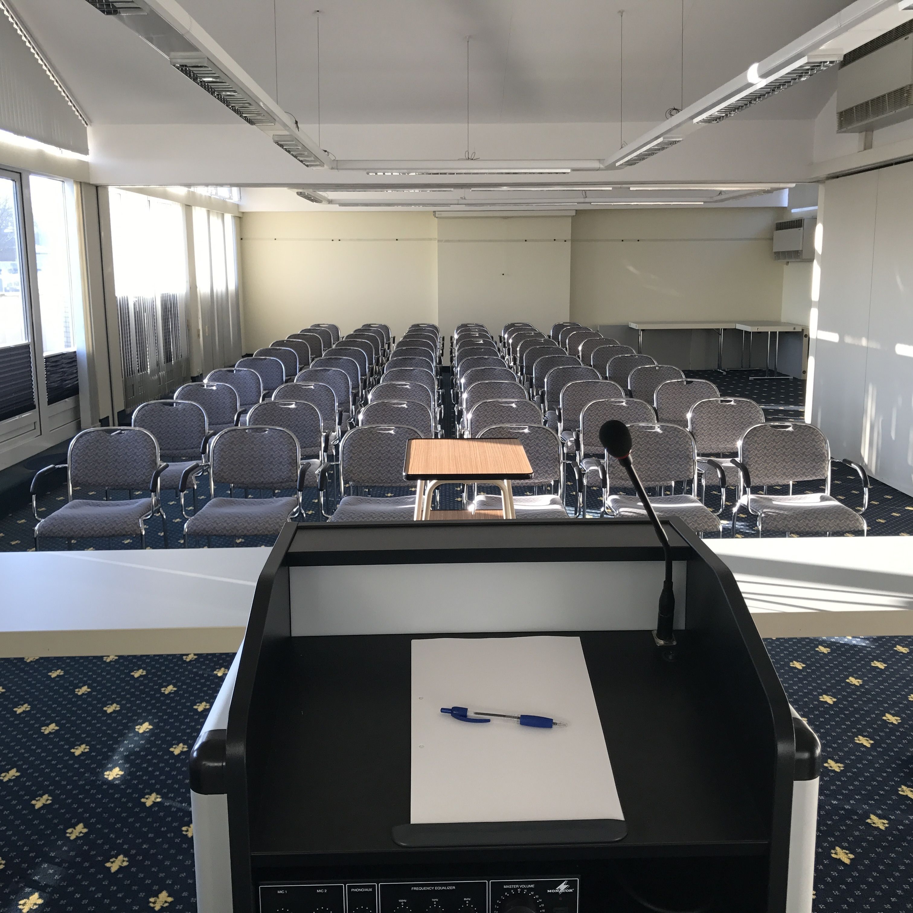 A conference room with rows of chairs, a podium with a microphone, and a table with a pen.