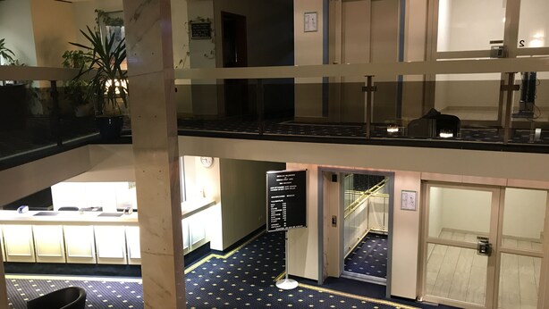 A multi-level hotel lobby with a check-in counter, a staircase, and a seating area with a potted plant. - Karaman Group Hotel (Bad Nenndorf)