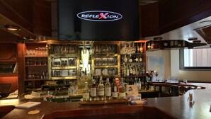 A bar area with a large screen displaying the Reflexion logo, shelves stocked with various bottles and glasses, and a counter with bar tools. - Karaman Group Hotel (Bad Nenndorf)
