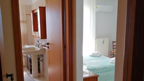 Un corridoio che conduce a un bagno con lavandino e specchio, e a una camera da letto con un letto e un comodino.