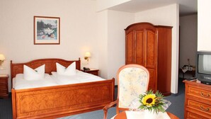Habitación de hotel con cama de madera, mesita de noche con lámpara, armario de madera, televisor y sillón estampado con motivos florales.