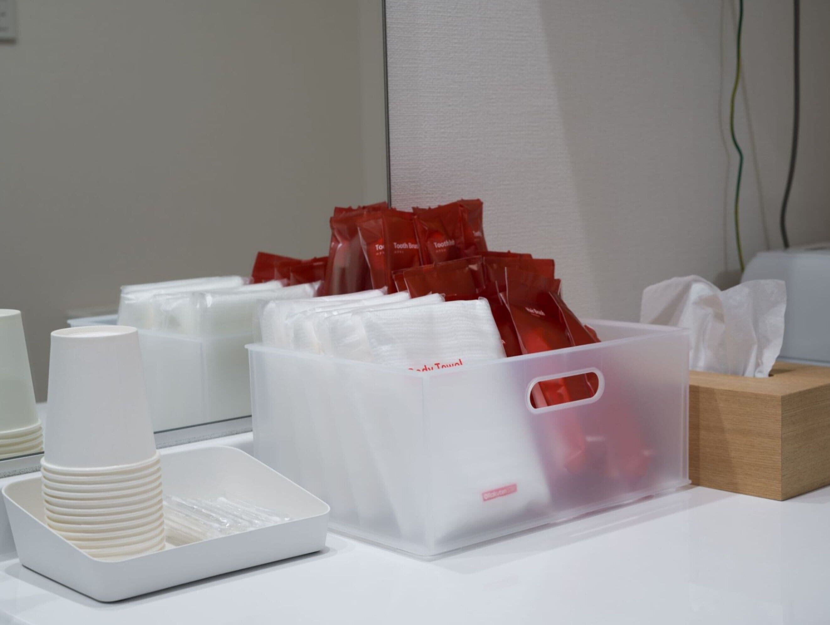 Une chambre d’hôtel avec une table blanche, une pile de tasses blanches, un récipient en plastique contenant des sachets rouges et une boîte en bois.