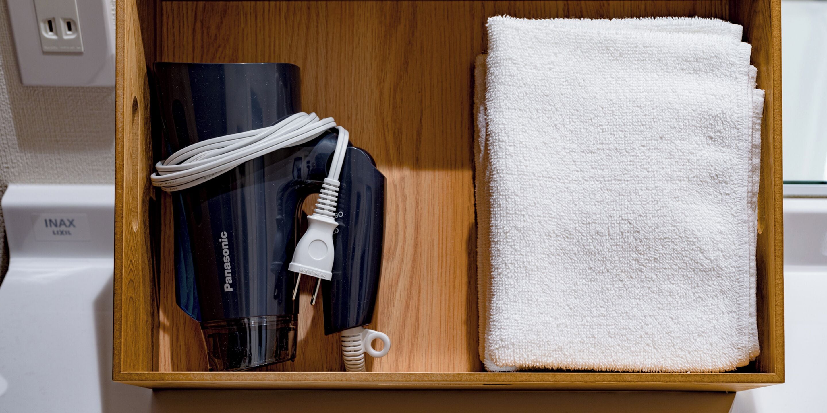 A wooden drawer containing a Panasonic hairdryer, a white towel, and a white charging cable.
