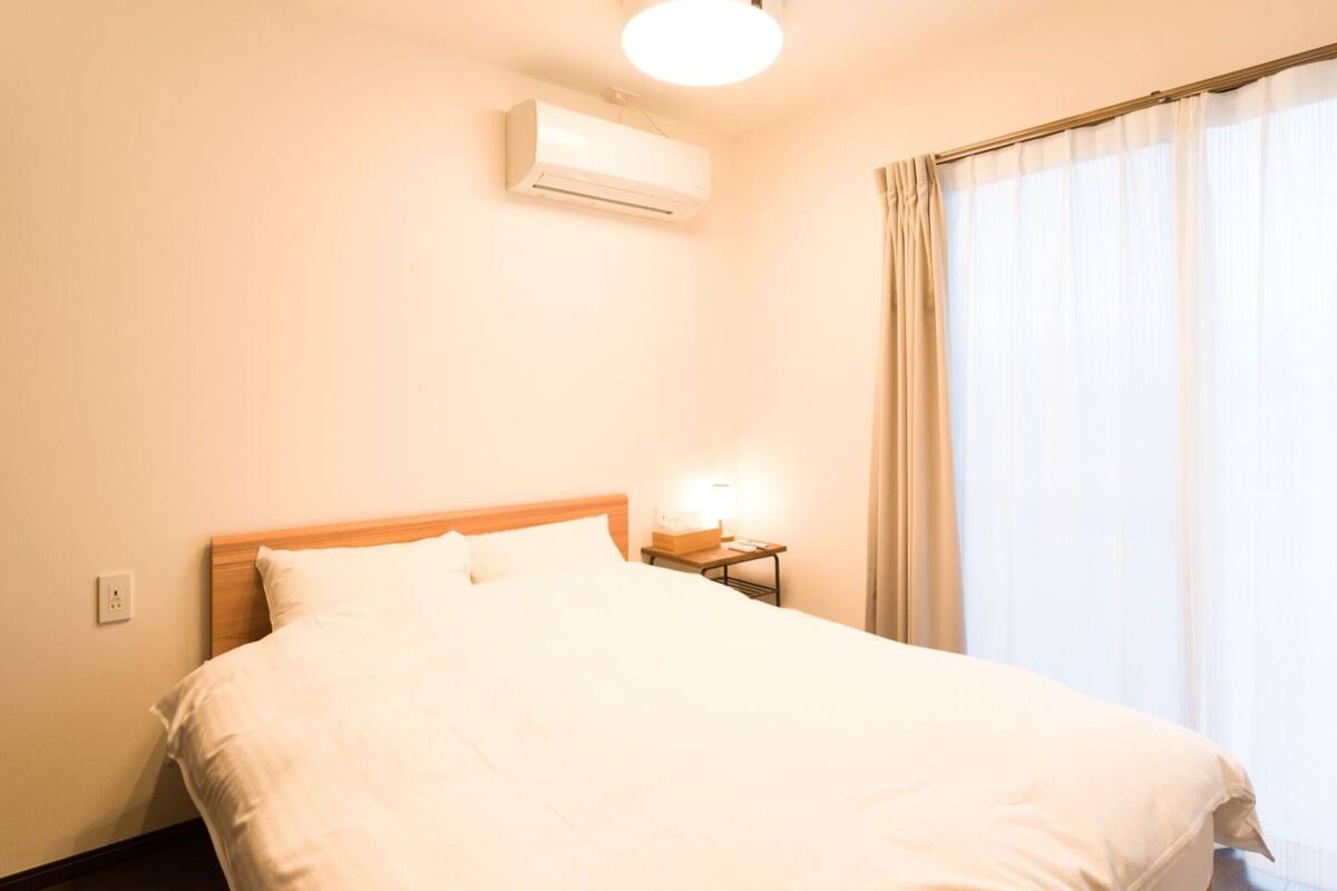 A hotel room with a bed, an air conditioning unit, a bedside table with a lamp, and a window with curtains.
