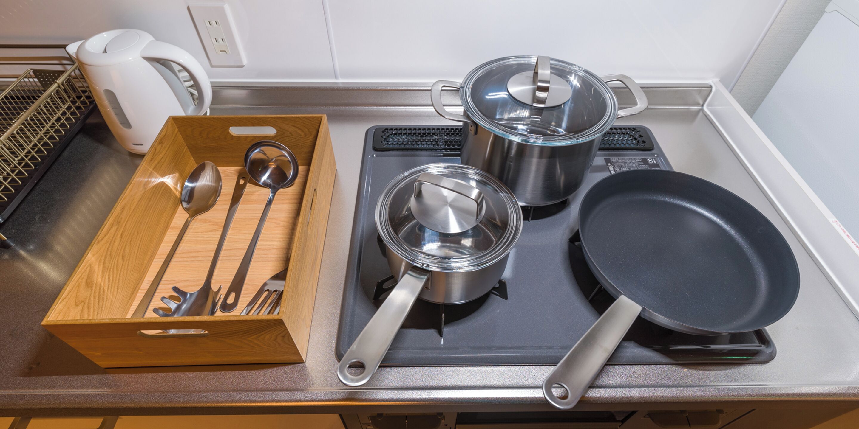 Un piano di lavoro in cucina con una scatola per utensili di legno, due pentole in acciaio inox con coperchi, una padella e un bollitore.