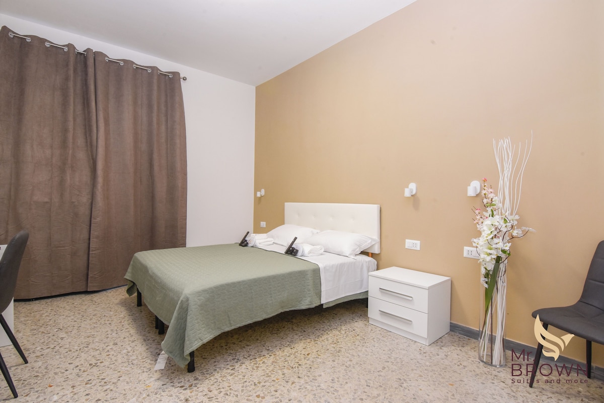 A bedroom with a bed, a nightstand, a chair, a desk, and a vase of flowers.