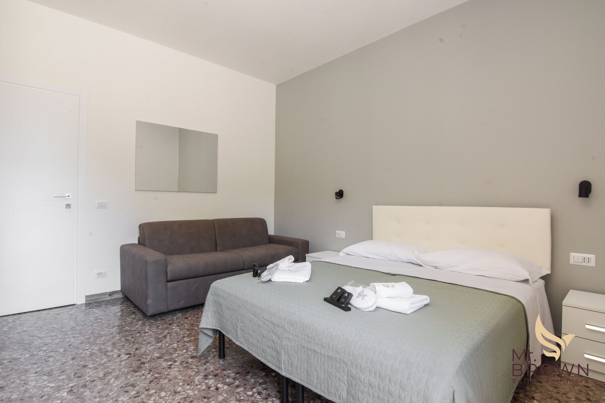 A hotel room with a bed, a grey sofa, a small table, and a mirror on the wall.