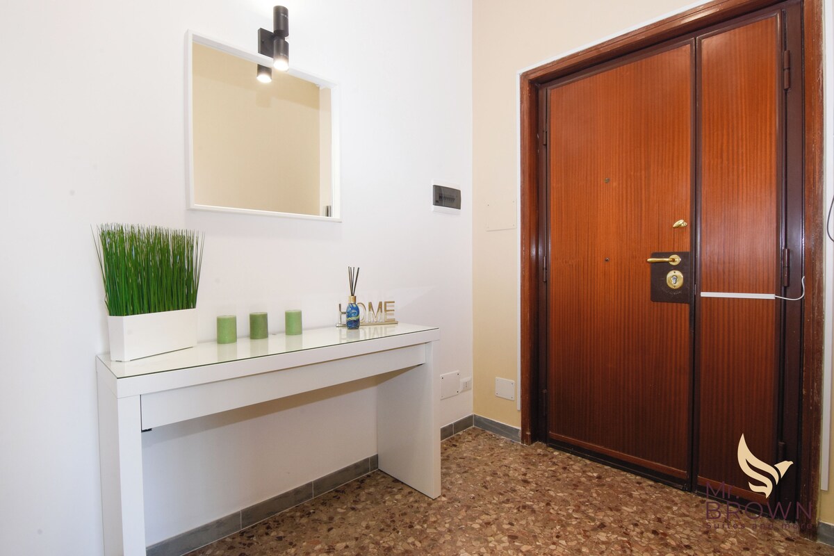 A white console table with a mirror above it, a potted plant, and a "HOME" sign. A wooden door with a brass handle is visible to the right.