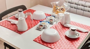 A dining table set with a white tablecloth, red placemats, a glass pitcher, a white teapot, a jar of Nutella, a bag of bread, and a white cup with saucer. - Guest House Mr Brown (Rome)