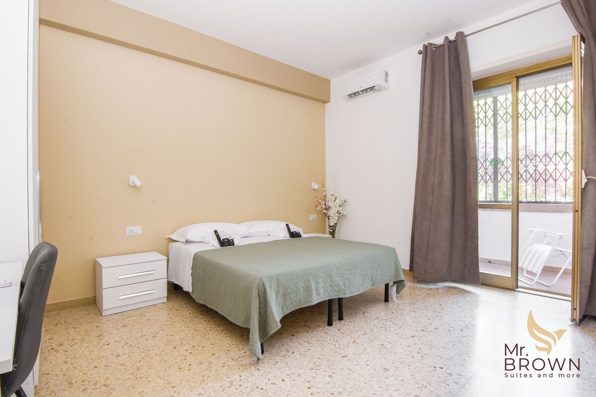 A bedroom with a bed, a nightstand, a chair, a window with curtains, and a view of the outside.