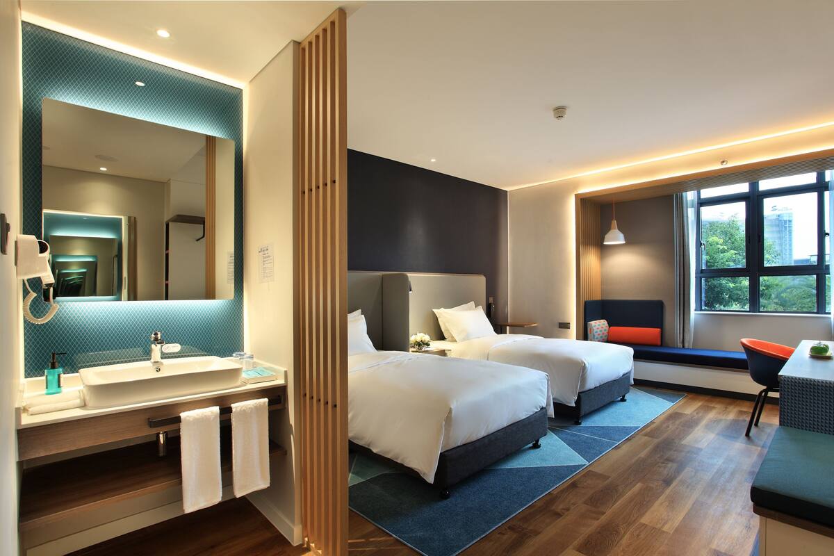 Une chambre d’hôtel moderne dotée d’un grand miroir, de deux lits, d’une salle de bain avec un lavabo et un porte-serviettes, et offrant une vue sur l’extérieur par une fenêtre.