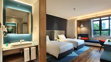 Une chambre d’hôtel moderne dotée d’un grand miroir, de deux lits, d’une salle de bain avec un lavabo et un porte-serviettes, et offrant une vue sur l’extérieur par une fenêtre.