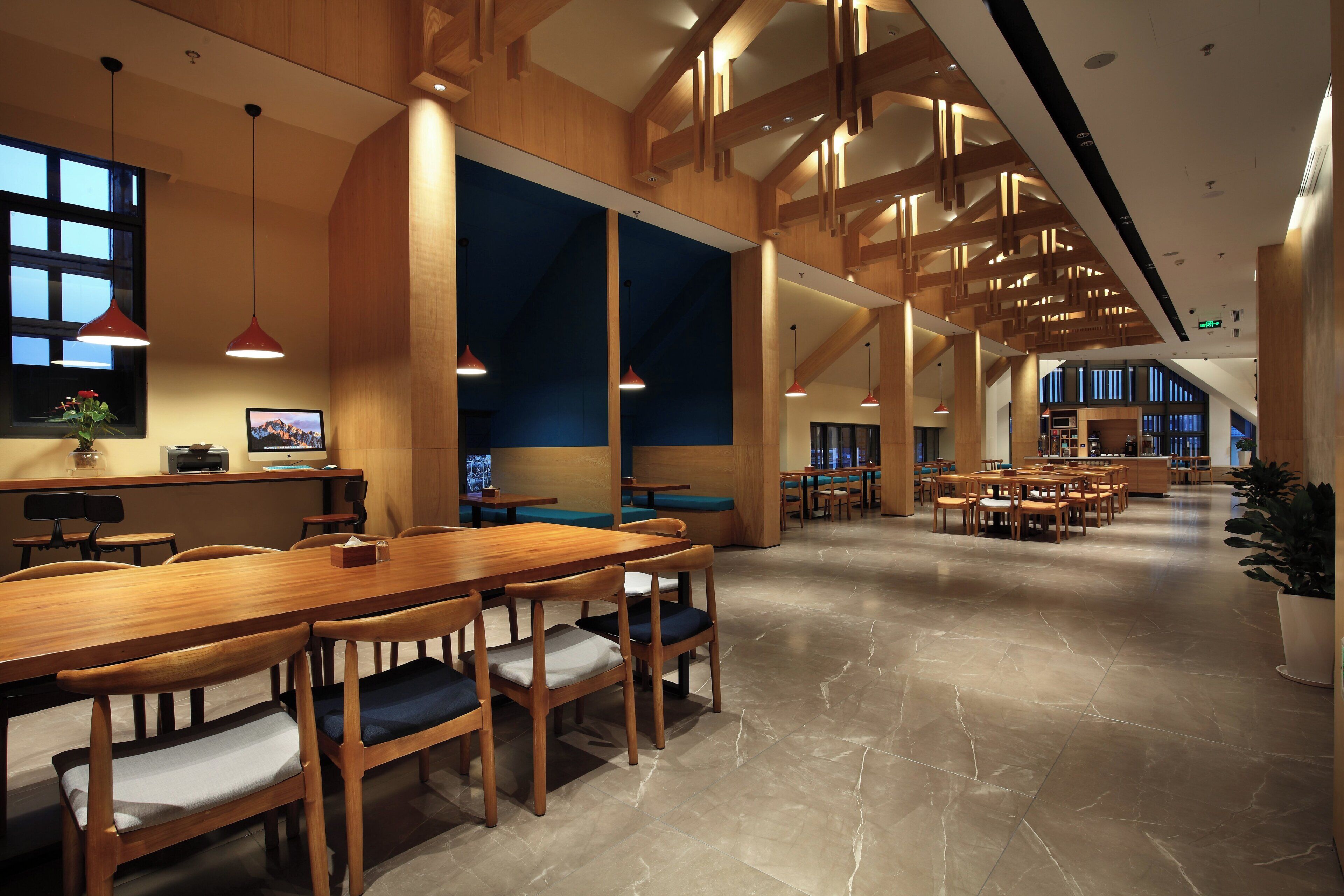 A spacious indoor area with a long wooden table and chairs, a large window, and a high ceiling with wooden beams.
