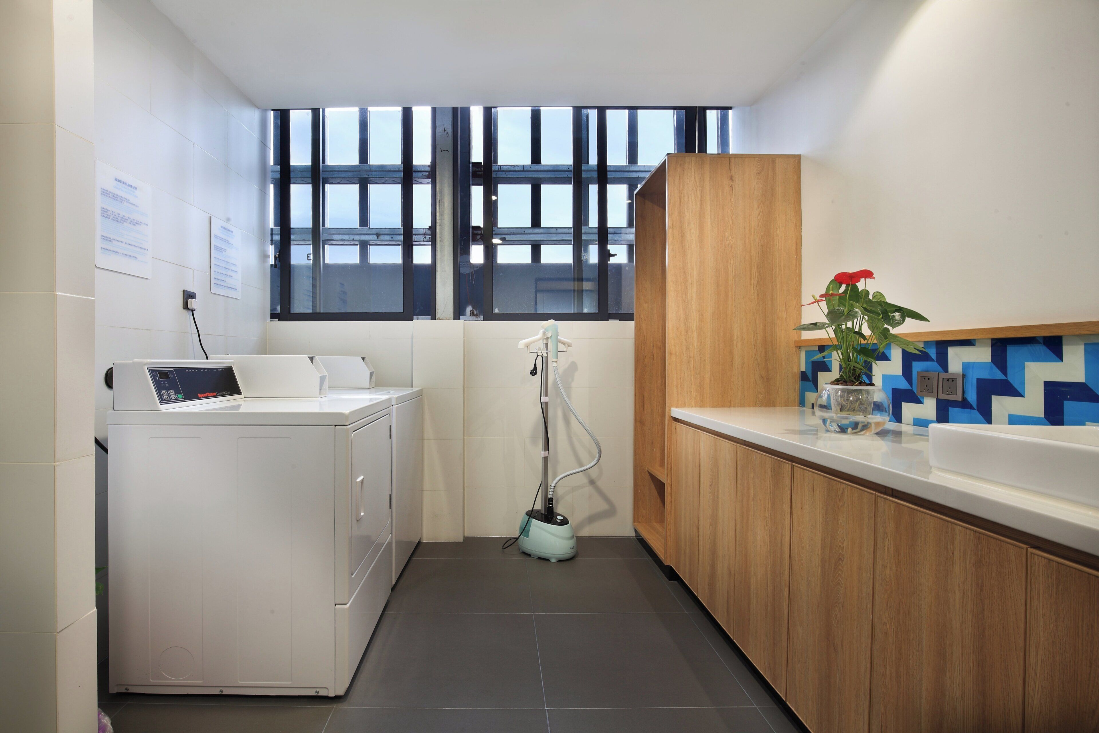A compact hotel bathroom with a washing machine, a hand dryer, a wooden cabinet, and a decorative plant.