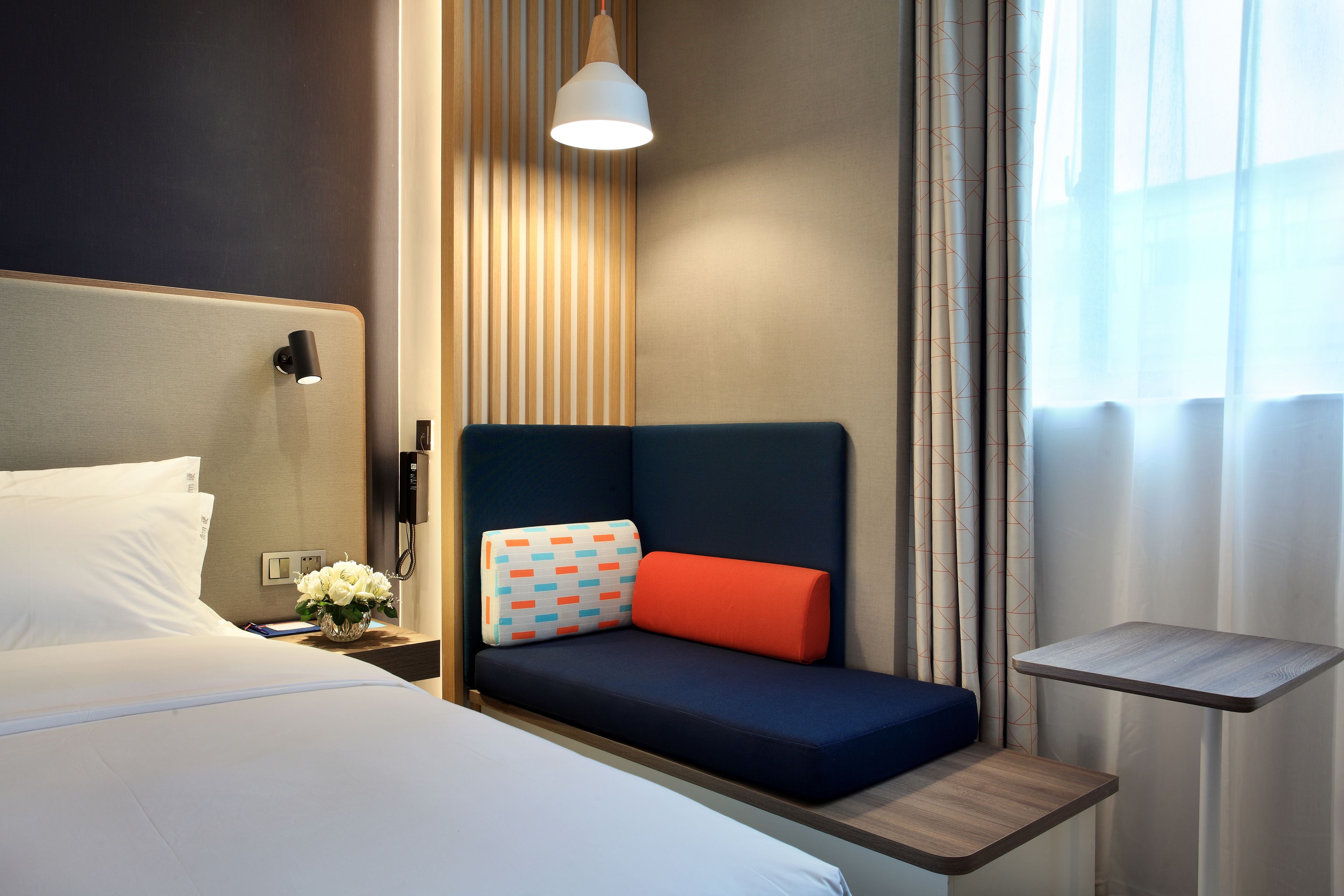 A hotel room with a bed, a bedside table with a lamp, a small seating area with cushions, and a view of the outdoors through sheer curtains.