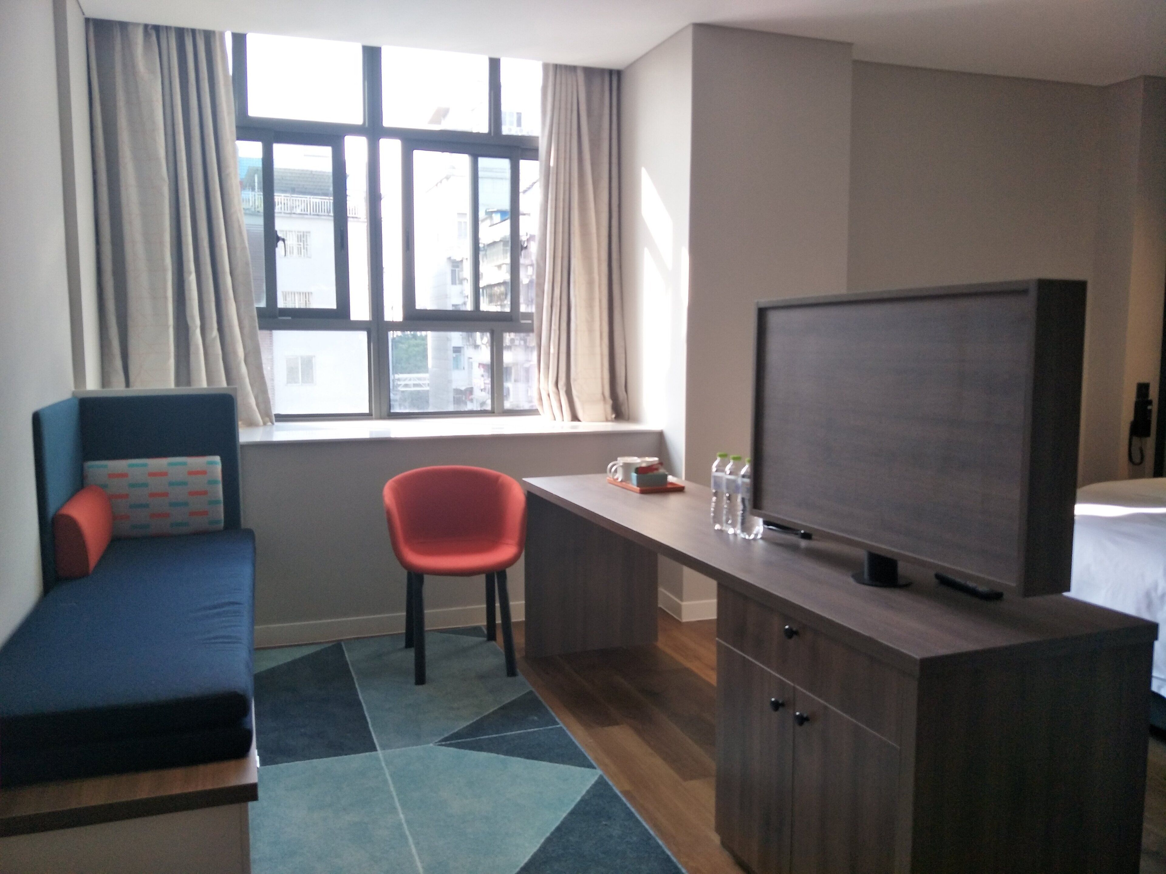 A hotel room with a bed, a desk with a television, a chair, and a window with curtains.