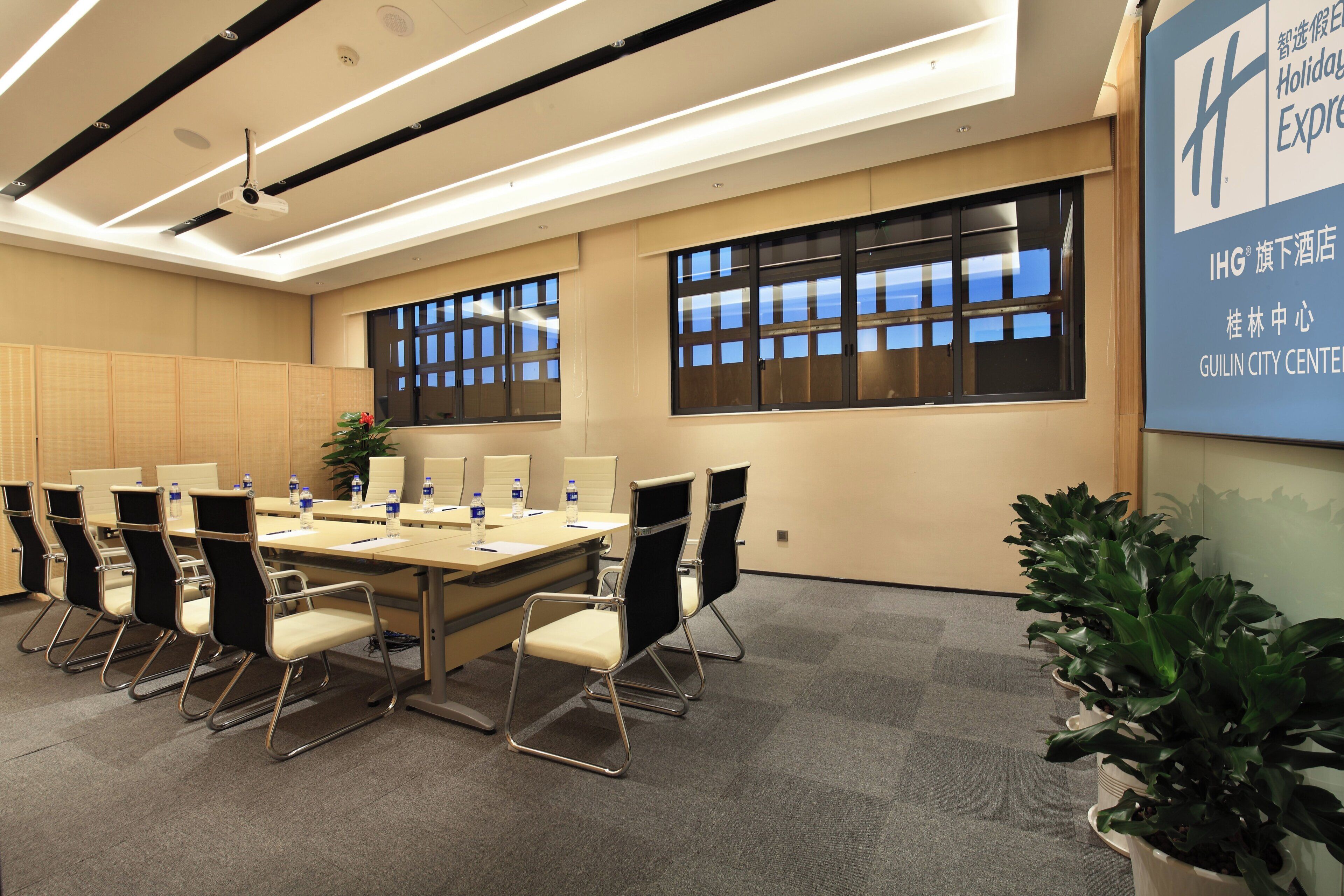 A conference room with a long table, chairs, and a projector. There are windows with blinds, a plant, and a sign on the wall.