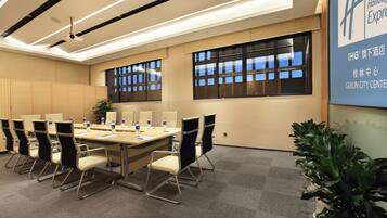 A conference room with a long table, chairs, and a projector. There are windows with blinds, a plant, and a sign on the wall.