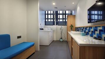 A modern laundry room with a blue sofa, washing machines, a vacuum cleaner, and a large TV mounted on the wall.