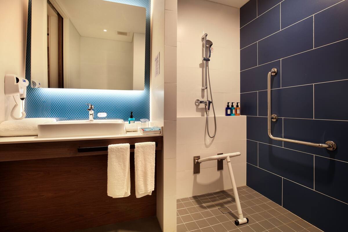 Une salle de bain équipée d’un lavabo, d’un miroir, d’un porte-serviettes, d’une douche et d’une barre d’appui.