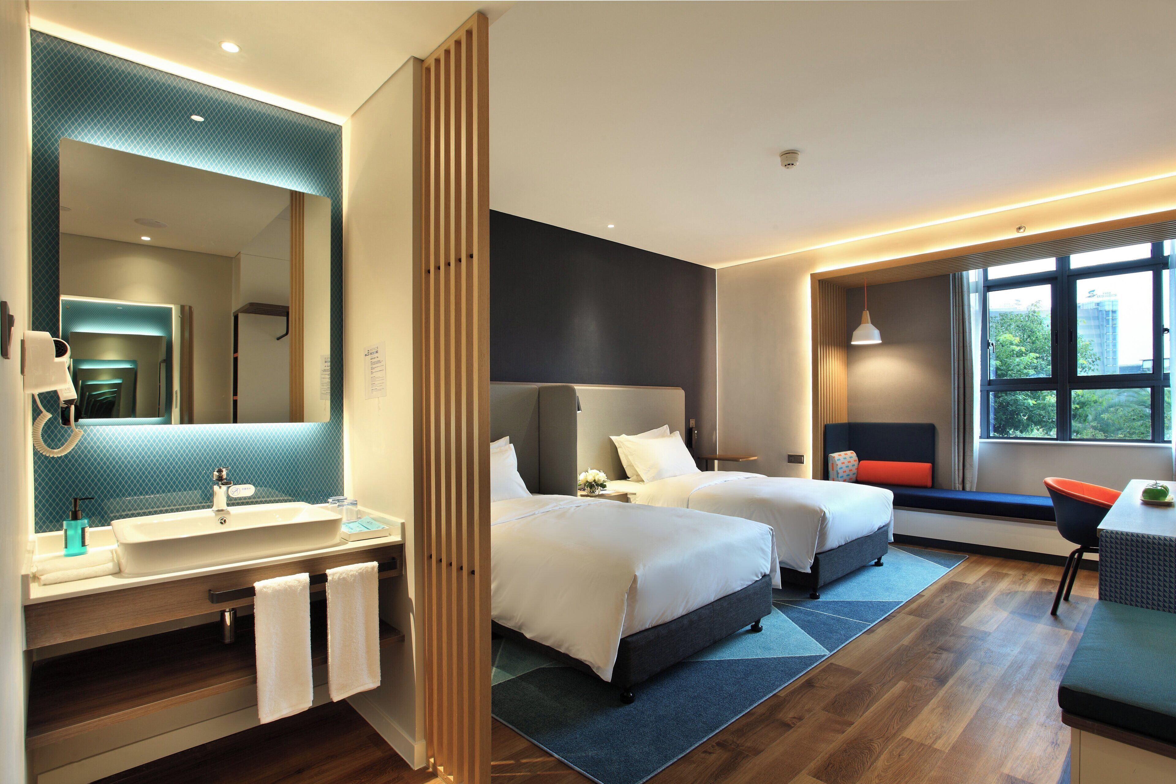 A hotel room with a large mirror, two beds, a bathroom with a sink, and a window with a view.