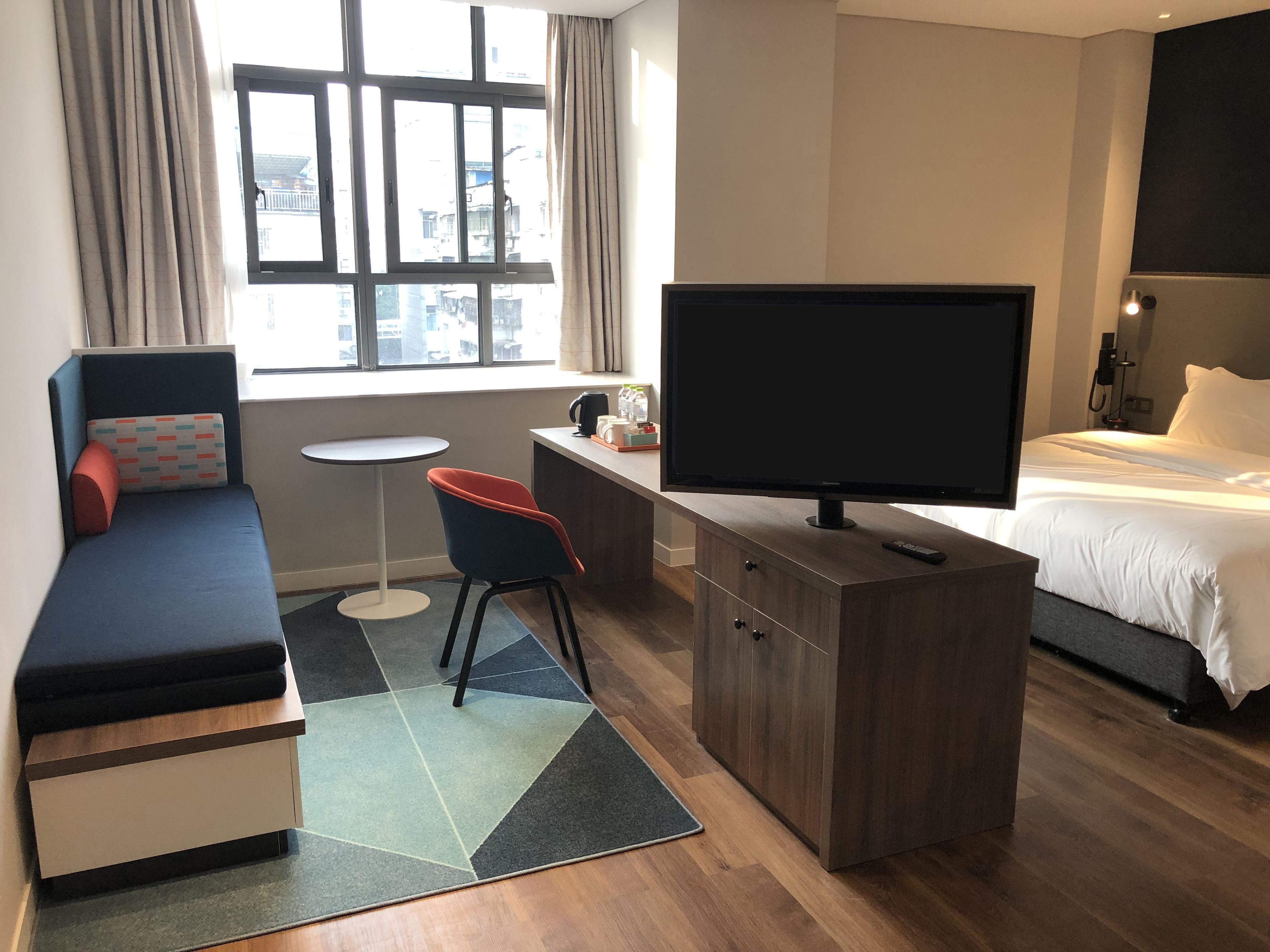 A hotel room with a bed, a TV, a desk with a chair, a sofa, and a window with curtains.