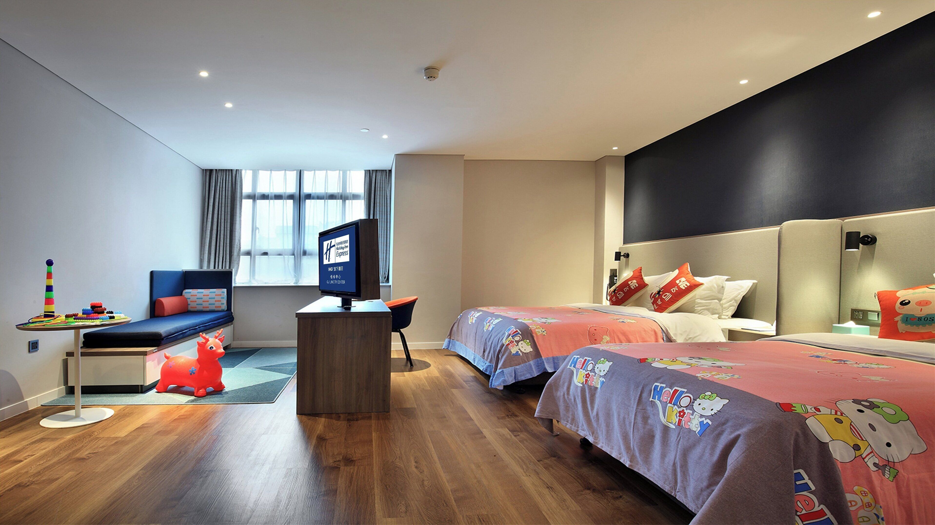 A modern hotel room with a wooden floor, a desk with a computer, two beds with Hello Kitty bedding, a window with curtains, and a view of the city.