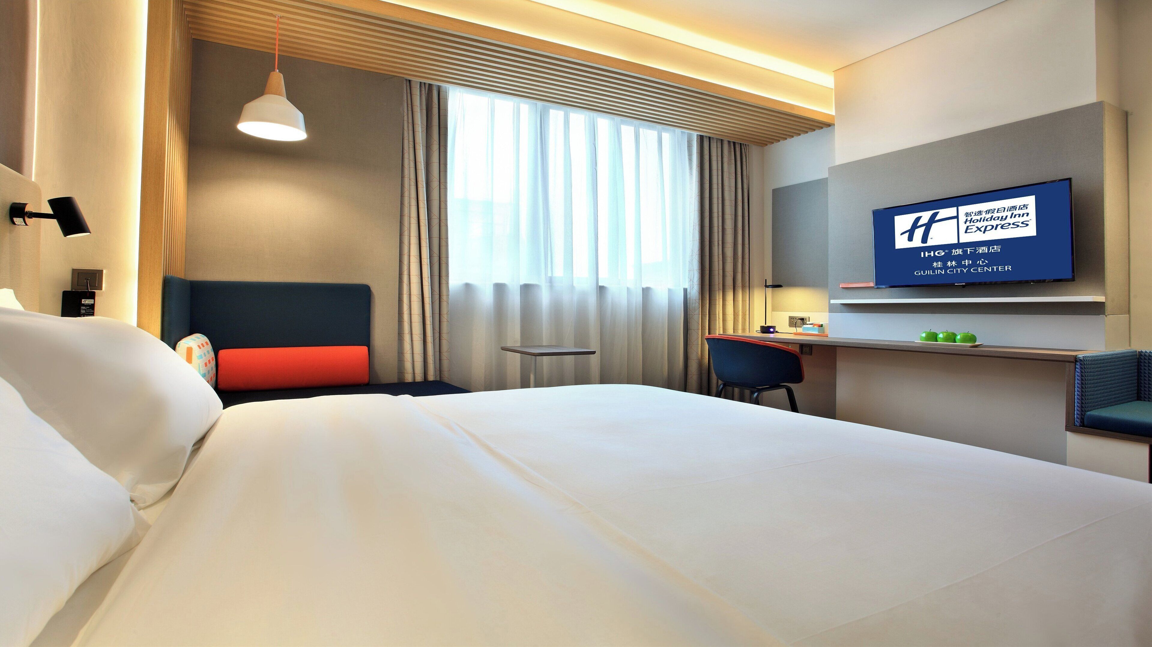 Habitación de hotel con cama, escritorio, silla, televisor y ventana con cortinas.