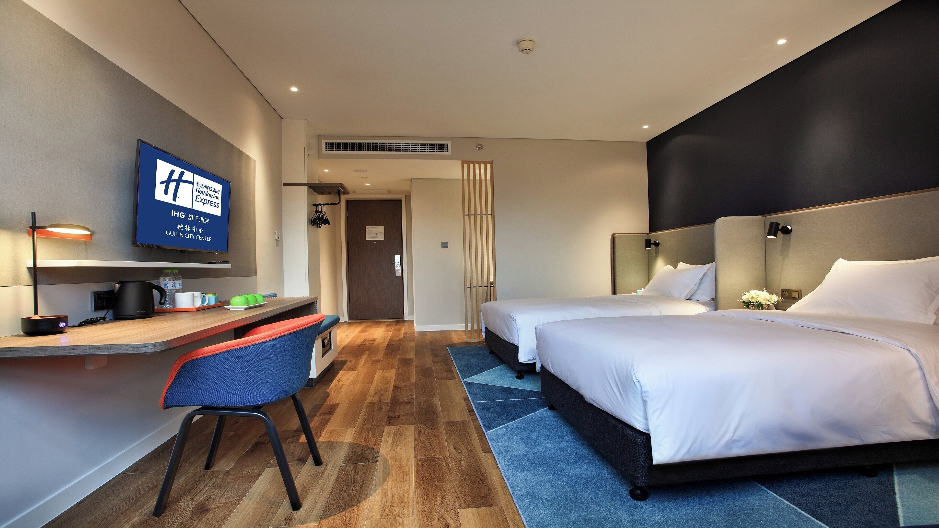 Une chambre d’hôtel avec un bureau, deux lits, une télévision et une porte de salle de bain.