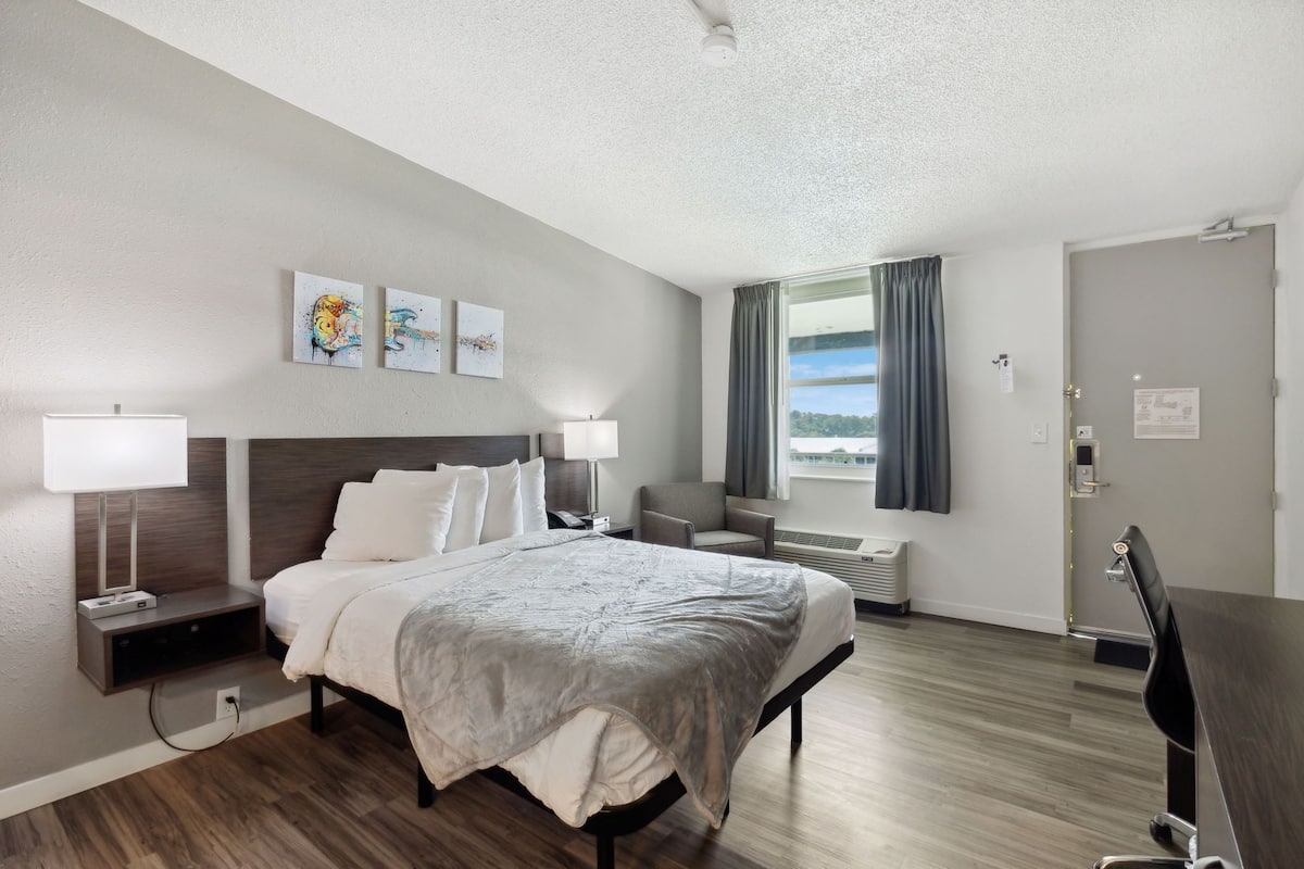 A hotel room with a bed, bedside lamps, a desk, a chair, a window with curtains, and artwork on the wall.