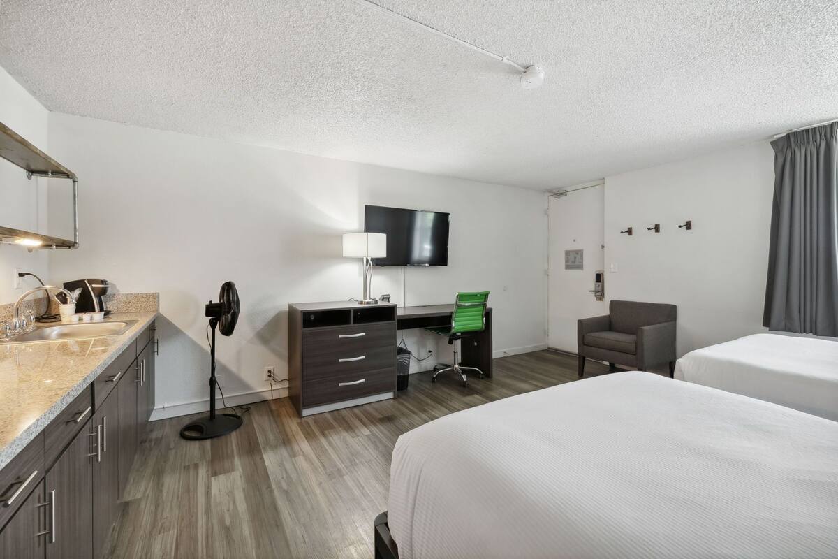 A hotel room with a bed, a desk with a chair, a TV, a fan, and a small seating area.