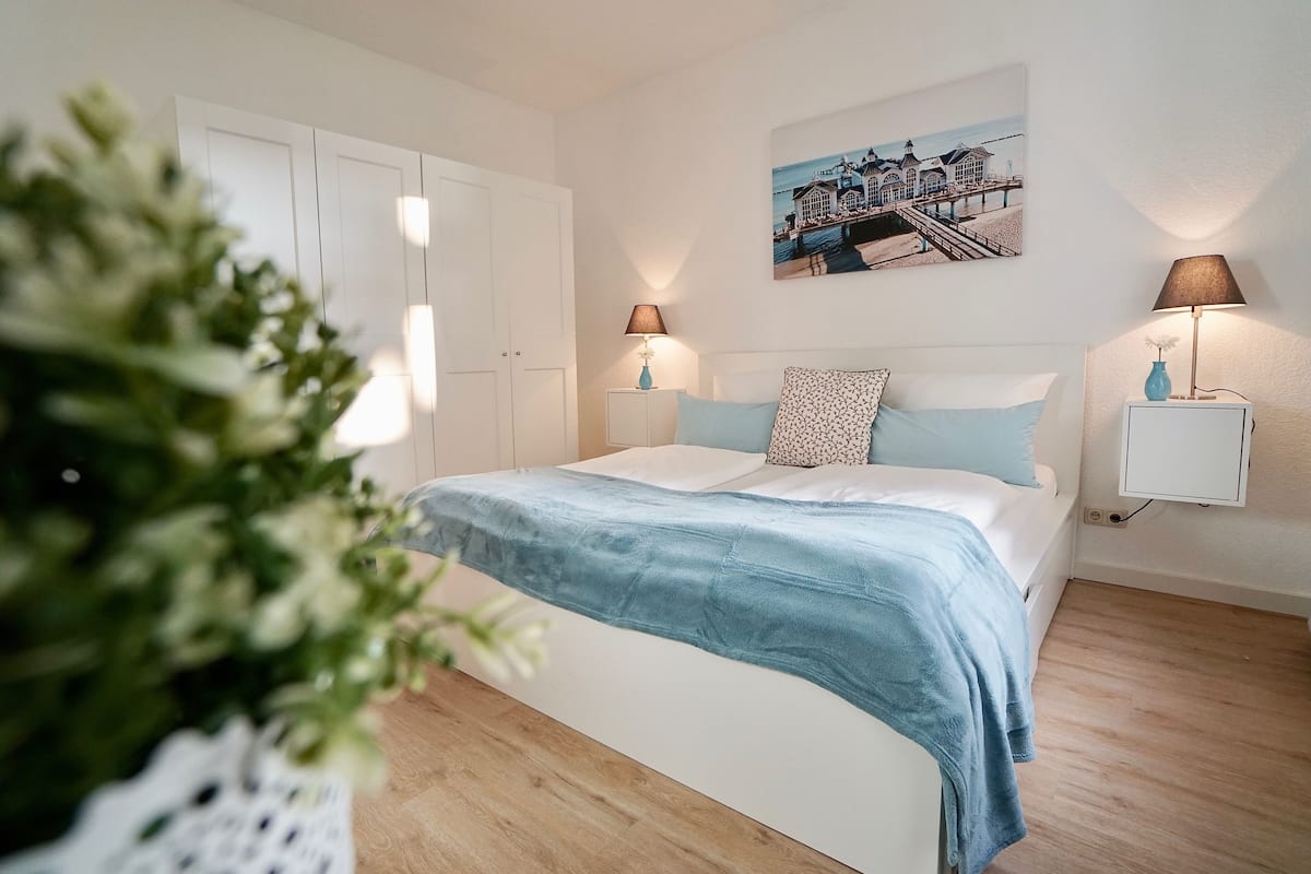 A bedroom with a bed, white wardrobe, two bedside tables with lamps, a wall-mounted picture, and a wooden floor.