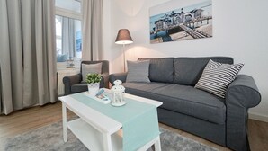 A modern living room with a grey sofa, a white coffee table, and a lamp. A painting of a coastal scene is on the wall. - Haus Johanneshorst (Sellin)