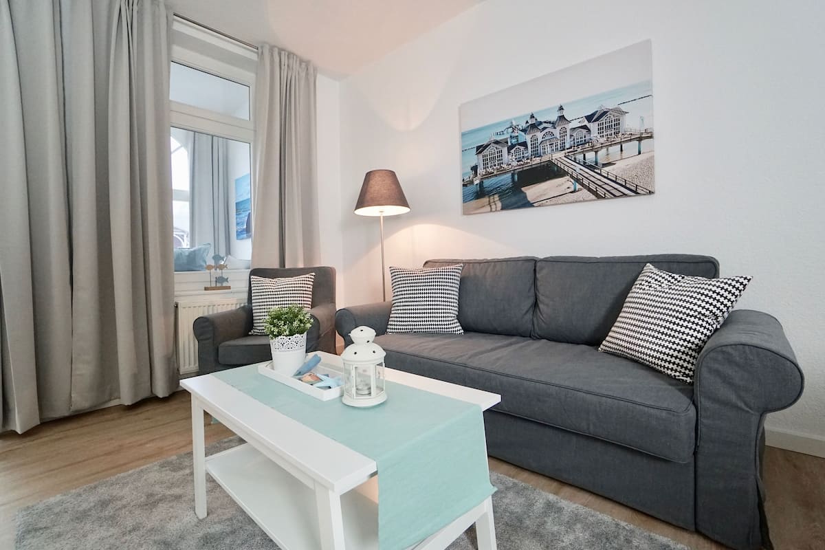 A modern living room with a grey sofa, a white coffee table, and a lamp. A painting of a coastal scene is on the wall.