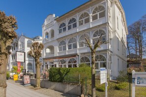 A white building with arched windows and a balcony, surrounded by trees and a paved walkway.