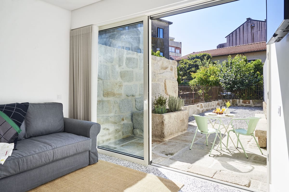 A modern living room with a grey sofa, a glass sliding door leading to an outdoor patio with a stone wall, and a view of neighboring buildings.