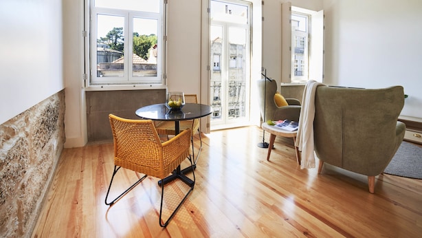 Een kamer met een houten vloer, een ronde tafel met stoelen, een bank en grote ramen met uitzicht op gebouwen en bomen.