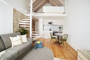 Un espacio habitable moderno con un sofá gris, una mesita redonda, una escalera de caracol y una zona de cocina.