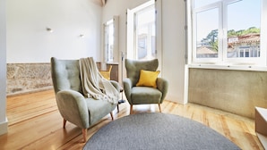 Een ruime kamer met een hoge plafond, houten vloer, een grijze fauteuil met een plaid, een groene fauteuil met een geel kussen en een groot raam met uitzicht op gebouwen en bomen.