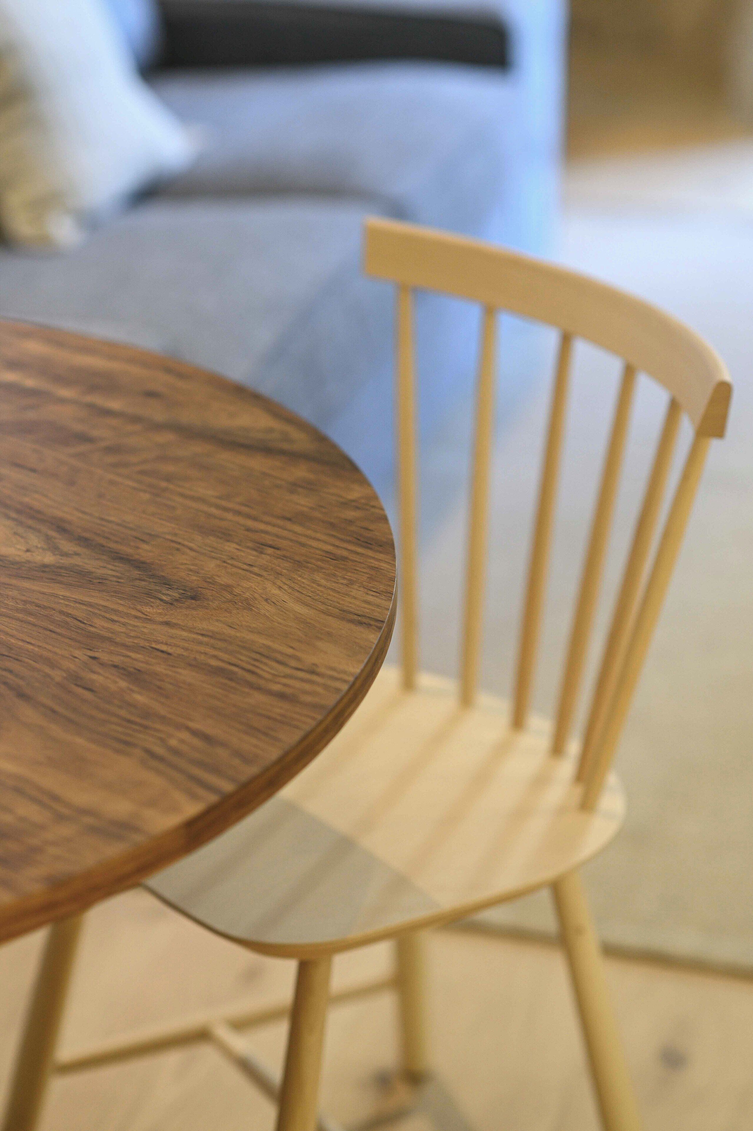A round wooden table with a glass top, positioned in front of a wooden chair with a high backrest.