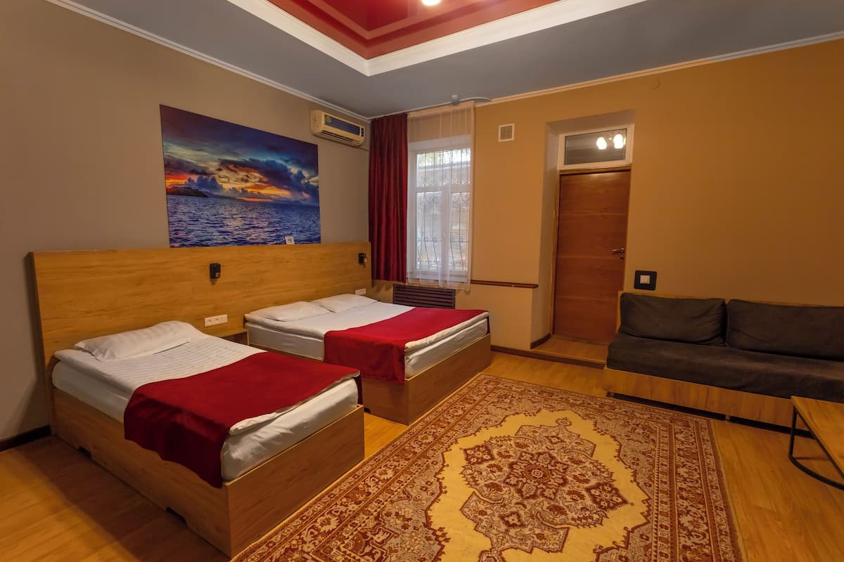 A hotel room with two wooden beds, a red blanket on one, a large rug, a sofa, a painting of a sunset, and a door with a window.