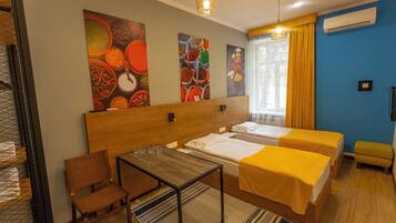 Quarto de hotel com cabeceira de madeira, duas camas individuais com colchas amarelas, uma mesa de madeira com copos, uma cadeira e uma obra de arte na parede.