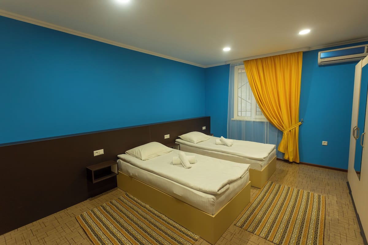 A hotel room with two beds, a window with yellow curtains, a small bedside table, and a patterned rug.